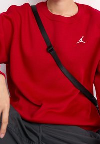 Person trägt einen roten Pullover mit weißem Jumpman-Logo und schwarzen Umhängeband, sitzt mit einer Hand auf dem Knie.