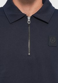 Close-up of a person wearing a navy blue zip-up shirt with a collar and a stitched rectangular logo patch on the chest.