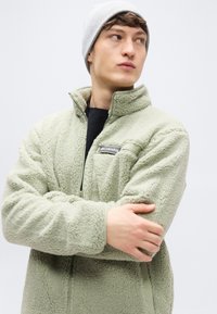 Hellgrüne Fleecejacke mit hohem Kragen und halbem Reißverschluss, ausgestattet mit einer linken Brusttasche und gerippten Bündchen. Kombiniert mit einer hellgrauen Mütze.