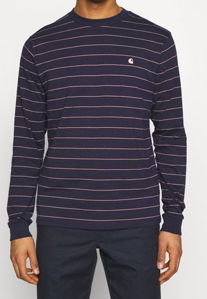 Persona que lleva una camisa de manga larga azul marino con finas rayas horizontales rosas y un pequeño logo rosa en el pecho izquierdo.