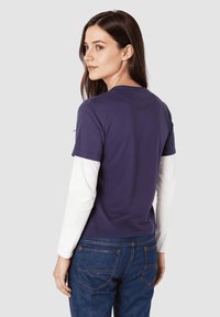 Camisa morada de manga corta superpuesta sobre una camiseta blanca de manga larga, combinada con jeans azules. Tejido suave, diseño sencillo y cuello redondo.
