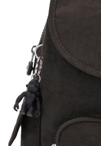 Zaino in tela nera con un flap arrotondato, dotato di una cerniera argentata e un piccolo portachiavi a forma di scimmia attaccato al tiretto della cerniera.