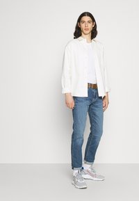 Camisa blanca de botones sobre una camiseta sin mangas blanca, jeans azules, cinturón marrón y zapatillas grises con acentos rojos, con un estilo casual.