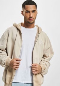 Beige zip-hoodie med en rummelig pasform, blød tekstur og en hvid logo detalje. Båret over en simpel hvid t-shirt og lyseblå denim.