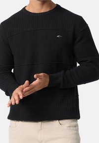 Pull noir avec tissu texturé et rayures horizontales. Poignets et ourlet côtelés. Petit logo sur la poitrine.