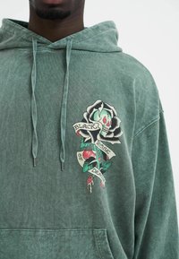 Groene hoodie van vervaagde stof met een grote afbeelding van een zwarte roos, een schedel en een banner met de tekst "Zwarte Roos Van Dood."