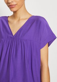 Lila V-ringad topp med korta ärmar, med ett plisserat mönster och mjukt material. Tyget verkar lätt och slätt.