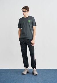 Mörkgrå sport-t-shirt med grön logotyp, kombinerad med svarta träningsbyxor med sidostripes och grå sneakers med texturerad detalj.