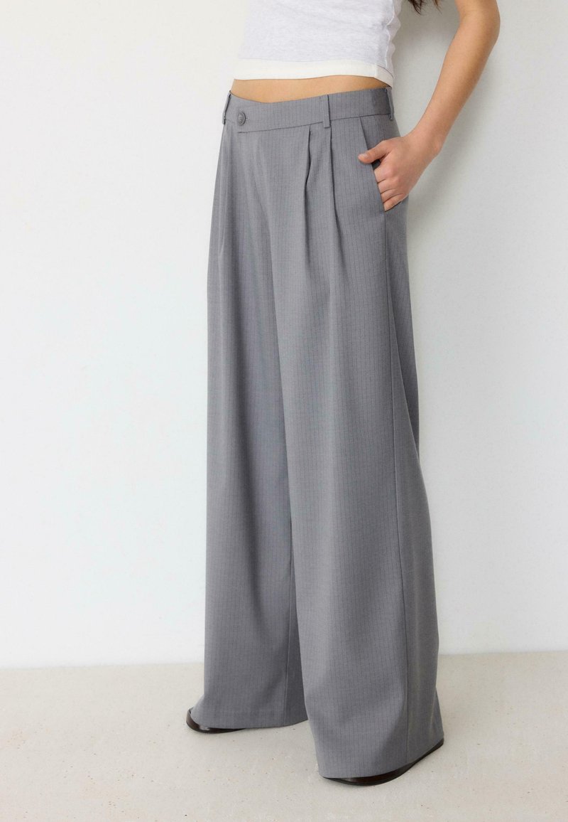 Femme vêtue d'un pantalon gris taille haute à jambes larges avec des plis et d'un top court blanc, main dans la poche, debout contre un mur blanc.