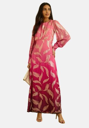 Vrouw in een lange roze ombre jurk met gouden veerpatronen, opgeblazen lange mouwen, houdend een gouden clutch, draagt gouden hakken.