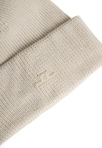 Stickad beige mössa med ribbad textur och vikkant, med en liten präglad logotyp på framsidan. Enkel och funktionell design.