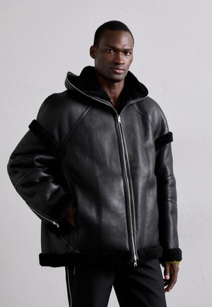 Schwarze Lederjacke mit einem mit Fell gefütterten Kragen, mit Reißverschluss vorne, seitlichen Taschen und schwarzen Fellakzenten an den Ärmeln und am Saum.