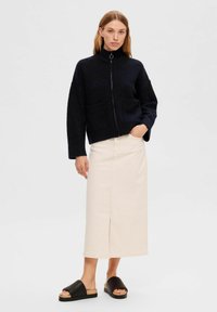 Marineblauwe zip-up sweater met hoge kraag en twee voorzakken, gecombineerd met een lange beige rok en zwarte slip-ons. Vlakke textuur en casual ontwerp.