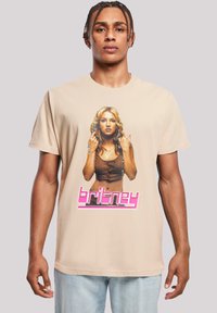 Béžové tričko s grafickým potiskem ženského portrétu a textem "britney" v růžovém kurzívním písmu pod ním. Krátký rukáv, ležérní design.