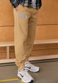 Καφέ φόρμες με το κείμενο "BOSTON" σε λευκό στο αριστερό τσεπάκι, διαθέτουν πλαϊνές τσέπες και ελαστικά τελειώματα. Συνδυάζονται με λευκά sneakers Nike.