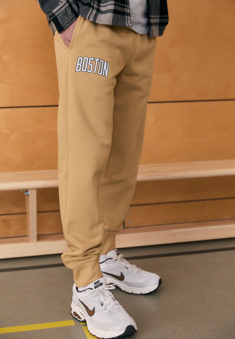 Καφέ φόρμες με το κείμενο "BOSTON" σε λευκό στο αριστερό τσεπάκι, διαθέτουν πλαϊνές τσέπες και ελαστικά τελειώματα. Συνδυάζονται με λευκά sneakers Nike.