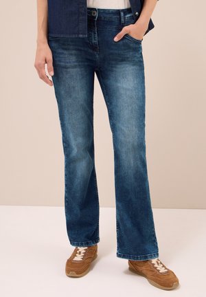 Persoon in blauwe bootcut jeans met één hand in de zak en bruine sneakers, staande voor een effen beige achtergrond.