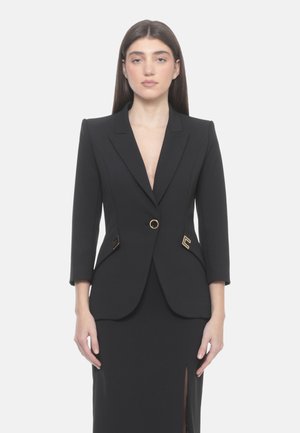 Donna con capelli lunghi e scuri indossa un blazer nero su misura con bottoni dorati e una gonna abbinata con spacco frontale, in piedi davanti a uno sfondo semplice.