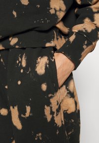 Sweat-shirt et pantalon tie-dye noir et beige avec poignets côtelés, une main partiellement dans la poche du pantalon.