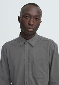 Chemise grise à manches longues avec un col boutonné, dotée d'une poche plaquée et de boutons violets, fabriquée à partir d'un tissu texturé.