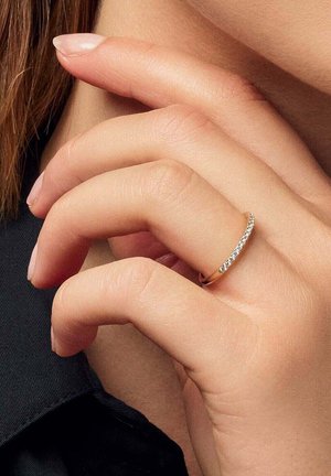 Hånd med lys hudtone iført en tynd guldbåndsring med små diamanter på ringfingeren, nær hagen og sort stof.