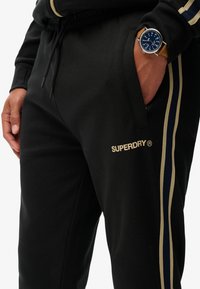 Man in zwarte Superdry trainingsbroek met beige en marineblauwe zijkanten, hand in zak, en een bruine leren horloge met een blauwe wijzerplaat.