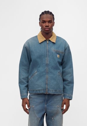 Jeune homme aux cheveux tressés portant une veste en jean bleue avec un col beige et un pantalon en jean assorti, debout devant un fond uni.