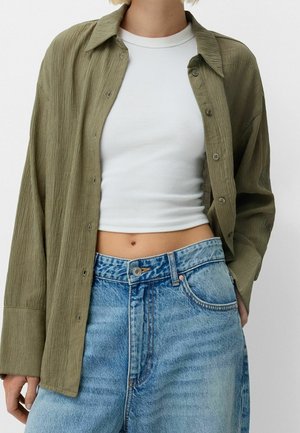 Jeune personne portant un crop top blanc sous une chemise texturée vert olive ouverte, associé à un jean en denim bleu clair taille haute.