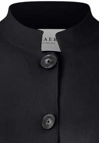 Černá texturovaná bunda s kulatým výstřihem a dvěma lesklými černými knoflíky zapnutými vpředu, s částečně viditelným štítkem uvnitř.