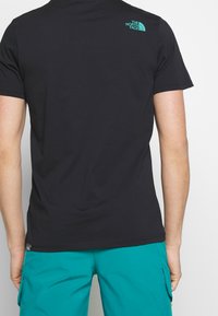 Svart bomull t-shirt med rund hals, med en turkos logotyp på den övre ryggen. Bärs tillsammans med teal cargo shorts.