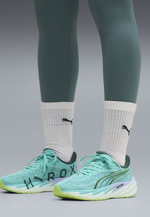 Jambes portant des leggings verts, des chaussettes blanches Puma et des chaussures de course Puma Nitro bleu clair avec semelles jaune-vert.