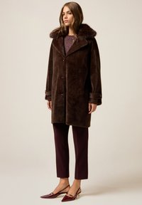 Kobieta ubrana w ciemnobrązowy płaszcz ze sztucznego futra, spodnie w kolorze bordowym oraz pasujące do nich bordowe szpilki na obcasie z odkrytą piętą, stoi na tle jednolitego jasnego tła.