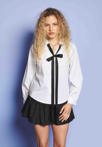 Νεαρή γυναίκα με σγουρά ξανθά μαλλιά, ντυμένη με μια λευκή μπλούζα με μαύρη κορδέλα και μαύρη φούστα, standing against a light blue background.