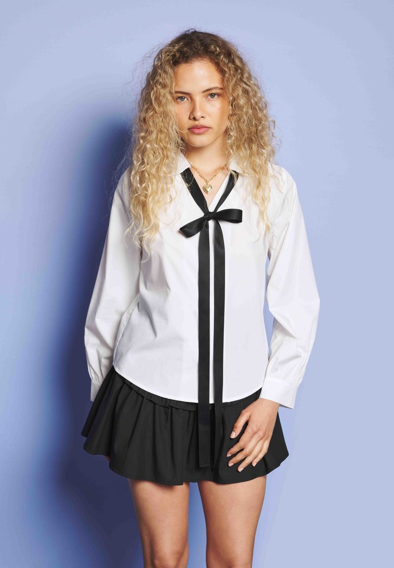 Νεαρή γυναίκα με σγουρά ξανθά μαλλιά, ντυμένη με μια λευκή μπλούζα με μαύρη κορδέλα και μαύρη φούστα, standing against a light blue background.