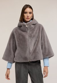 Szara peleryna z sztucznego futra z kapturem, z przednim zamkiem błyskawicznym, szerokimi rękawami i miękką teksturą, noszona na teksturowanej niebieskiej koszuli i spodniami z kordurowej tkaniny.