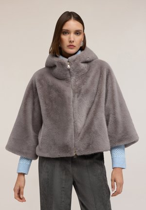 Grijze nepbont capuchoncape met een ritssluiting aan de voorkant, wijde mouwen en een zachte textuur, gedragen over een gestructureerd blauwe shirt en corduroy broek.