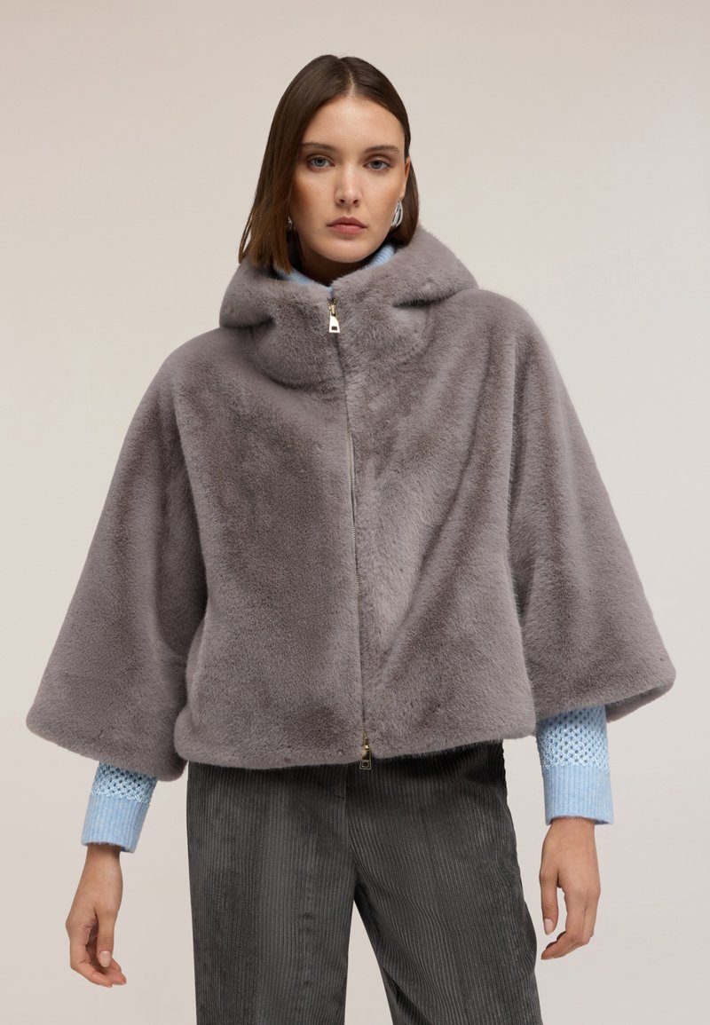 Szara peleryna z sztucznego futra z kapturem, z przednim zamkiem błyskawicznym, szerokimi rękawami i miękką teksturą, noszona na teksturowanej niebieskiej koszuli i spodniami z kordurowej tkaniny.