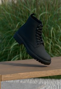 Stivale nero alla caviglia con punta rotonda, tomaia in pelle opaca liscia, robusta chiusura con lacci e suola in gomma spessa con texture a righe.