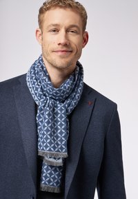 Blå mønstret tørklæde i en klassisk vævning, med frynsekant, båret over en mørk jakke, der fremviser indviklede geometriske mønstre.