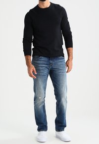 Mann steht mit einem schwarzen Langarmshirt, blauen, ausgewaschenen Jeans mit einem kleinen Riss am linken Oberschenkel und weißen Turnschuhen vor weißem Hintergrund.