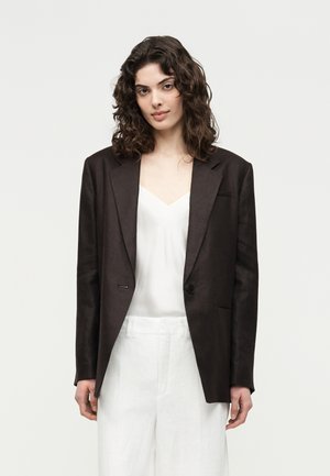 Frau mit lockigem dunklem Haar, die einen schwarzen übergroßen Blazer über einem weißen V-Ausschnitt-Oberteil und weißen weiten Hosen vor einem einfarbigen Hintergrund trägt.