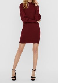Femme portant une robe tricotée ajustée bordeaux avec de longues manches bouffantes et des bottines à plateforme avec bride noire, se tenant contre un fond blanc.
