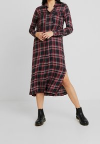 Abito camicia a quadri neri e rossi con bottoni frontali, due tasche sul petto, maniche lunghe e spacco laterale all'orlo. Indossato con stivali neri alla caviglia.