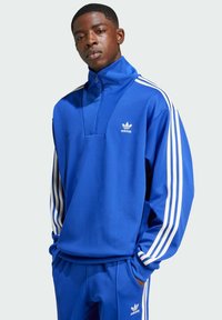 Blauer Quarter-Zip-Pullover mit weißen drei Streifen an den Ärmeln, mit einem Adidas-Logo auf der Brust und einem lockeren Schnitt.