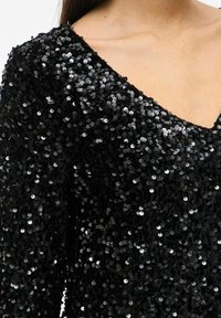 Juoda V formos iškirptės suknelė, padengta tankiai išdėstytais blizgančiais juodais blizgučiais, atidengiančiais viršutinę krūtinės ir pečių sritį.