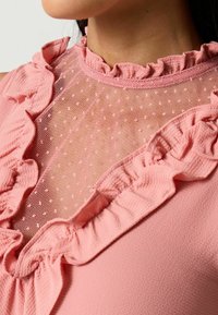 Růžová blůza s polka dot síťovinou nahoře, zdobená volánky a vysokým výstřihem, zdůrazňující jemnou texturu a ženské designové prvky.