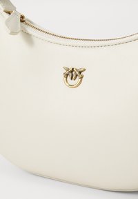Crème-kleurige handtas met een gestructureerd oppervlak, voorzien van een gouden metalen accent in de vorm van bladeren en een cirkelvormig detail aan de voorkant.
