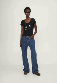 Μαύρο κοντομάνικο πουκάμισο με λογότυπο; μπλε φαρδιά τζιν παντελόνια; μαύρα σανδάλια; το μοντέλο στέκεται σε ελαφρύ φόντο, με τα χέρια χαλαρά στα πλάγια.