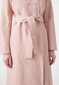 Vaaleanpunainen trenssitakki, jossa on kaksirivinen malli ja sileä kangas. Takissa on solmittava vyö ja näkyvät nappiyksityiskohdat.