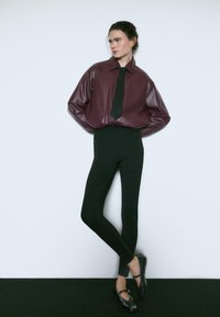Camicia over-size in finta pelle martora bordeaux con cravatta nera, abbinata a leggings neri aderenti e ballerine nere. Il modello è in piedi contro uno sfondo neutro.